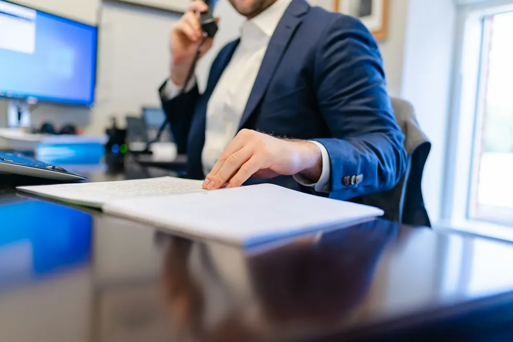 Professional in a suit on a phone call at his office desk, reviewing notes