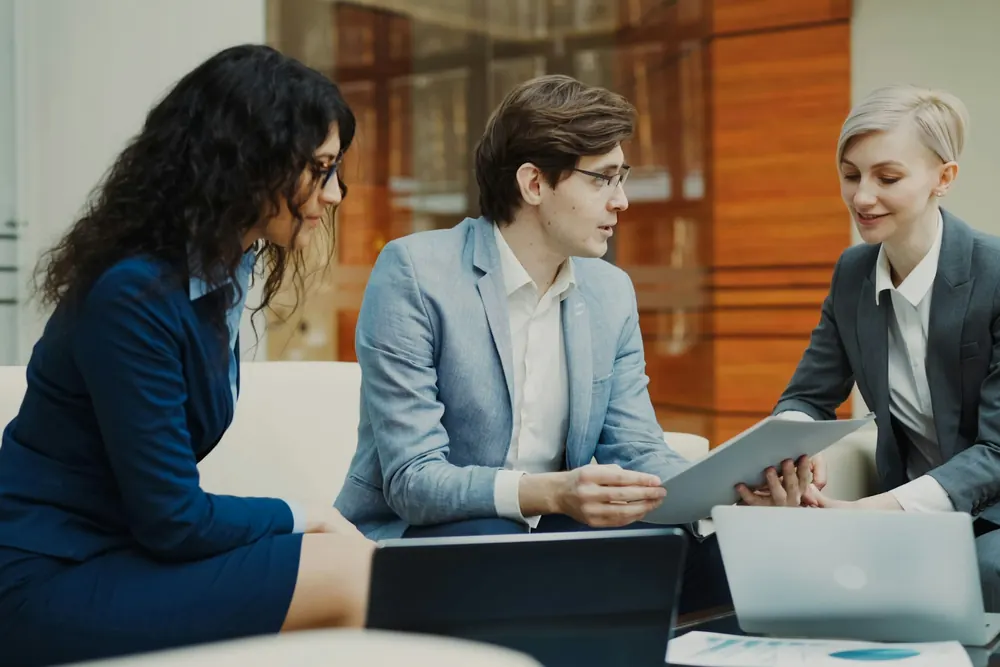 Three colleagues in business attire reviewing documents together in an office lounge